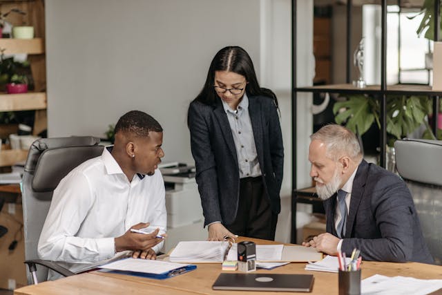 Illustrative image A Man in Black Suit Jacket Sitting at a Desk Beside a Man and a Woman used in Accordemy®'s training on Supervision Skills for Managers in Land/Portfolio Management